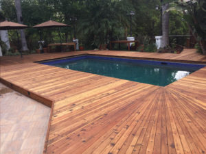 A well preserved and maintained redwood deck surrounding a swimming pool with thatched roofed cabanas.